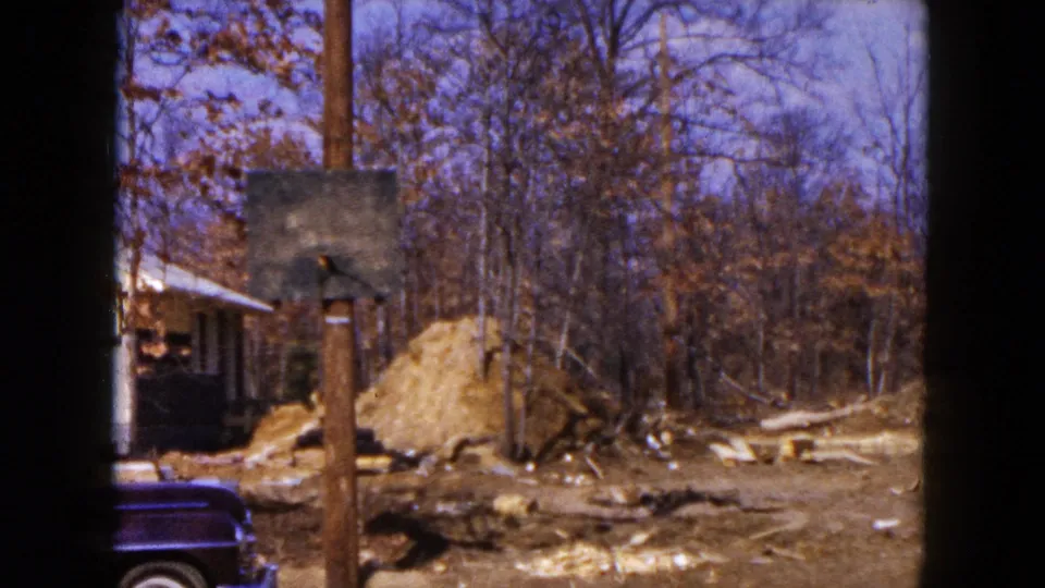 Hicksville, New York frame from 1954 showing a residential lot with dirt piles, trees, a utility pole, and a partial dark sedan.