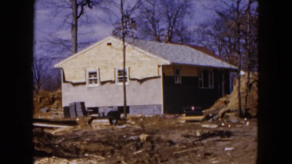1954 Hicksville frame showing a single-story house under construction with a bare yard, utility pole, dirt pile, and materials near the foundation.