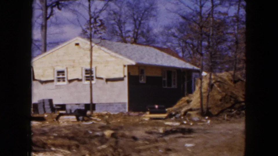 Hicksville 1954 home-movie frame showing a single-story house on a rough construction lot with dirt piles and bare trees.
