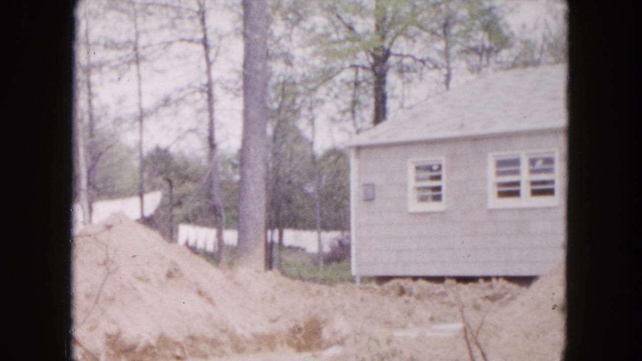 1954 Hicksville home-movie frame showing a modest light-colored house in a wooded lot with excavated sandy soil in front.
