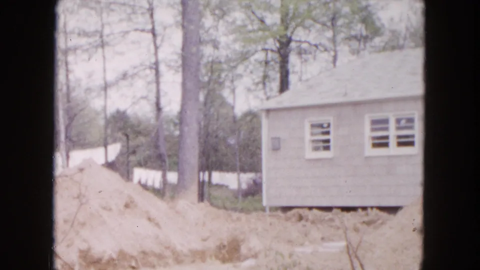 1954 Hicksville home-movie frame showing a modest light-colored house in a wooded lot with excavated sandy soil in front.
