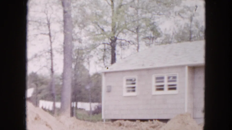 1954 Hicksville frame of a shingled house exterior with windows, a brick chimney, trees, and unfinished ground around the lot.