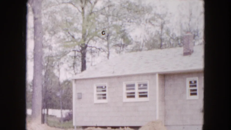 1954 Hicksville home-movie view of the house facade, showing windows, a chimney, nearby trees, and unfinished ground below.
