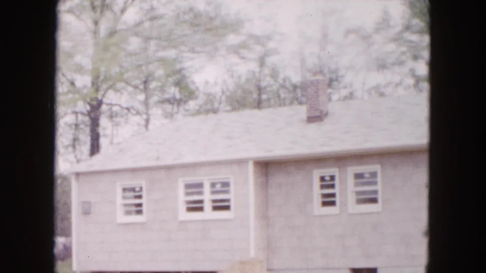 1954 Hicksville frame showing the side of a modest house in a wooded setting, with windows, chimney, and pale disturbed soil visible.