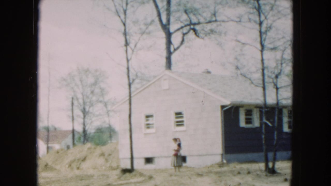 1954 Hicksville home-movie frame showing a small light-and-dark house, bare trees, uneven yard, and a woman near the wall.