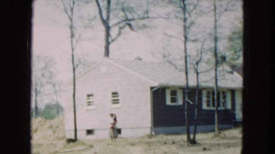 Hicksville, New York residential exterior from 1954 with a woman near a small house and tall bare trees in the yard.