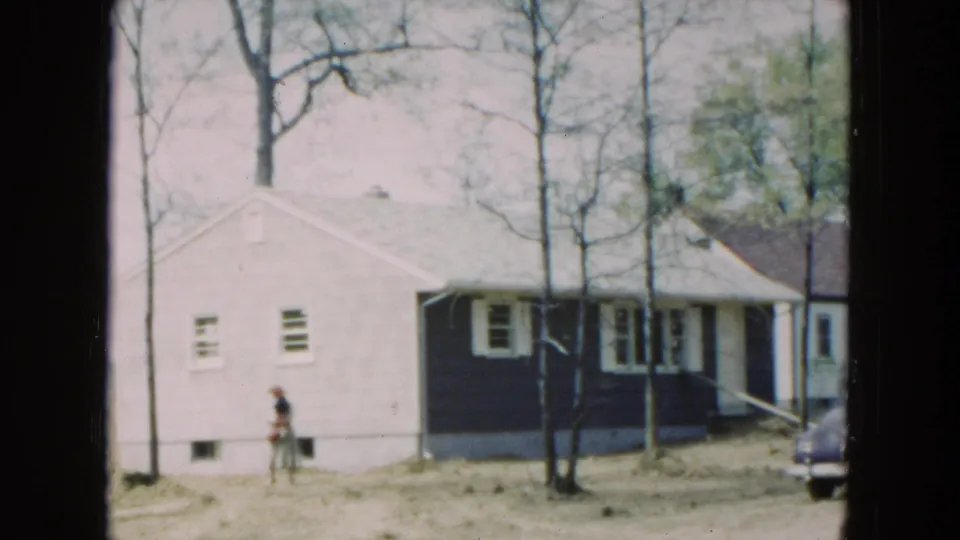 1954 8mm frame of a woman near a Hicksville house, with bare trees, uneven ground, and a neighboring residence in view.