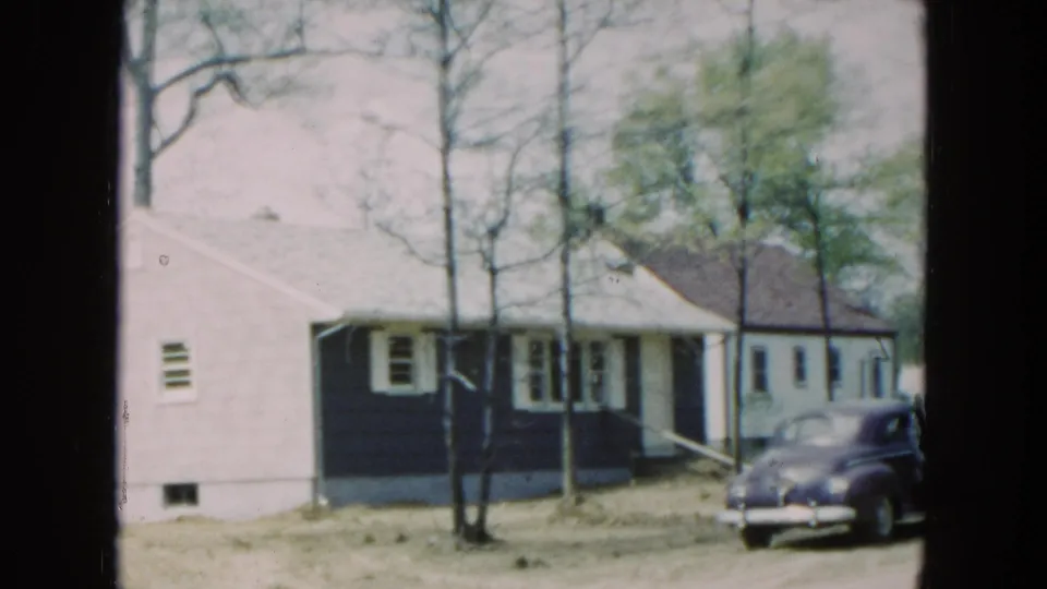 1954 Hicksville residential scene with a small house, leafless trees, uneven yard, and a rounded parked car at frame right.