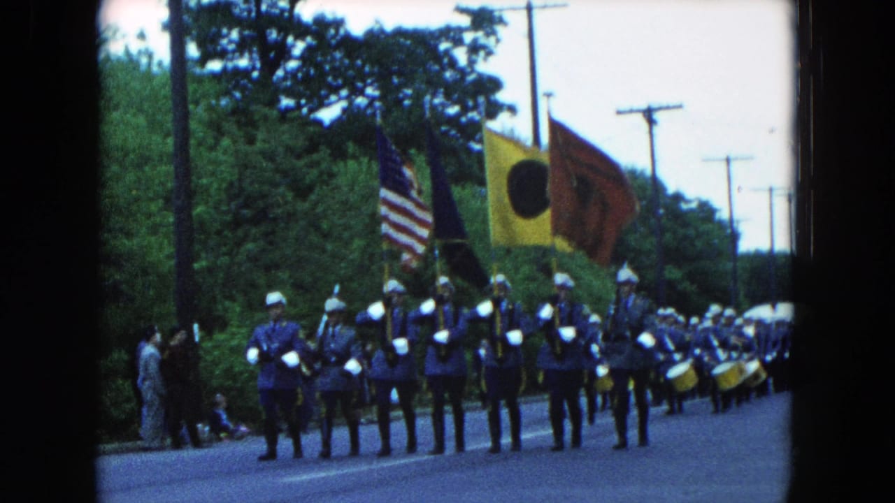 1954 Hicksville parade scene with uniformed marchers, white helmets, drums, and flags moving down a tree-lined road.