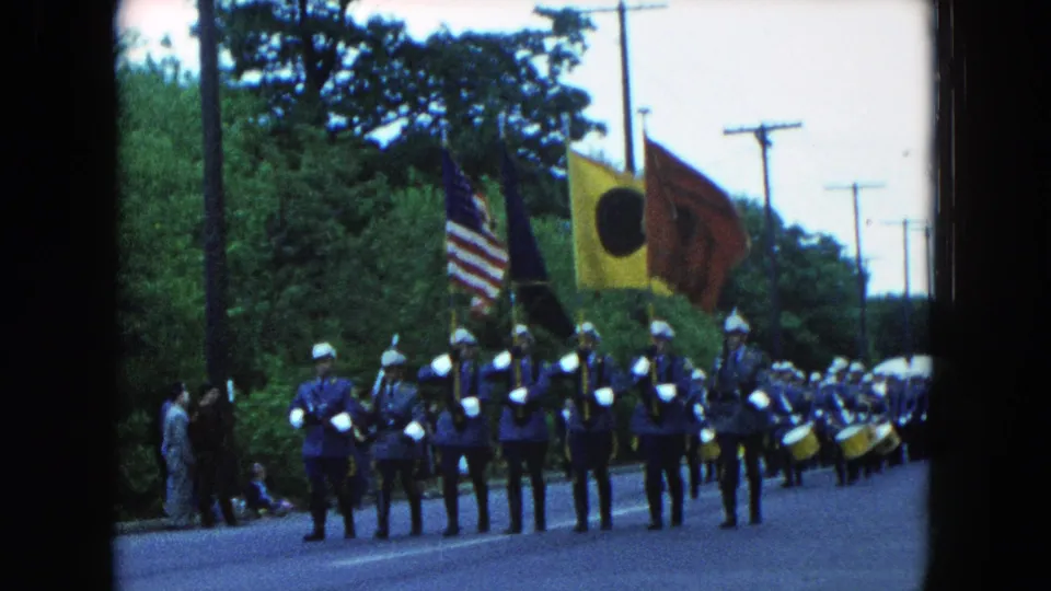 1954 Hicksville parade scene with uniformed marchers, white helmets, drums, and flags moving down a tree-lined road.