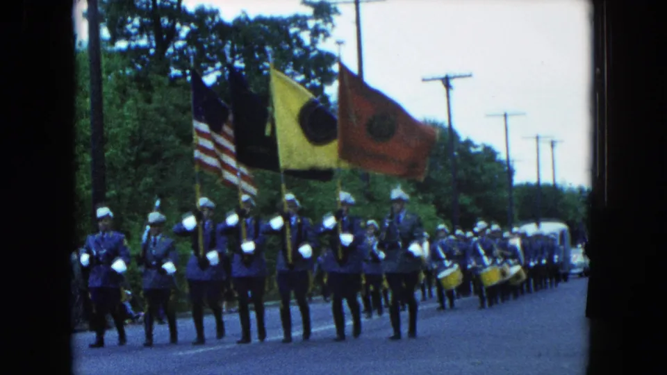 Uniformed band members and flag bearers march in a 1954 Hicksville patriotic parade, with spectators near the roadside.