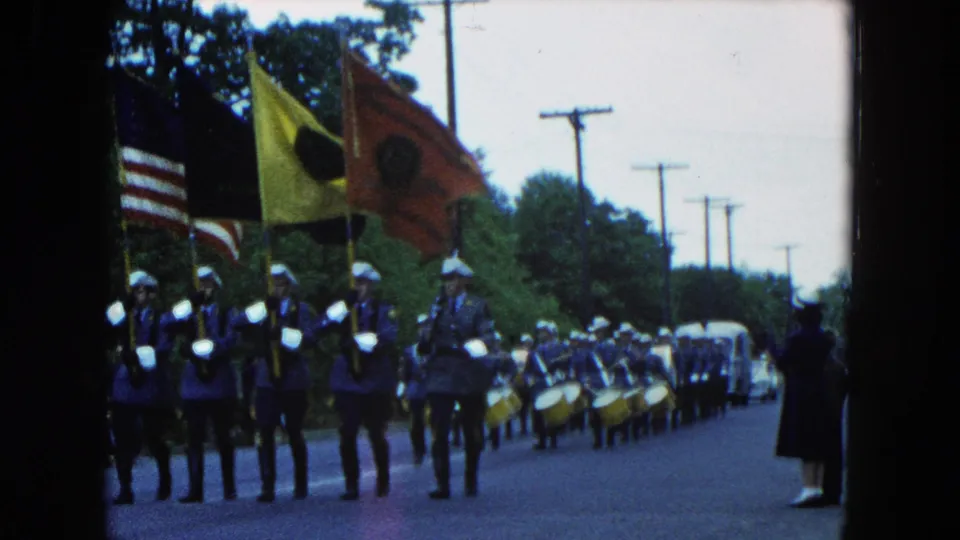 Roadside home-movie view of the Hicksville parade as flag bearers and drummers pass close to the camera.