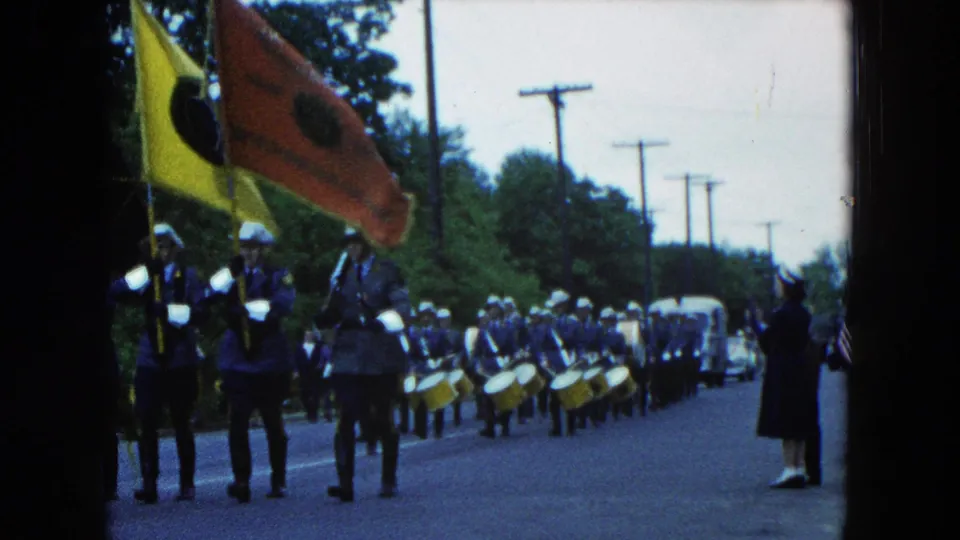 Uniformed marchers with large flags and snare drums continue through Hicksville as a spectator stands at the road edge.