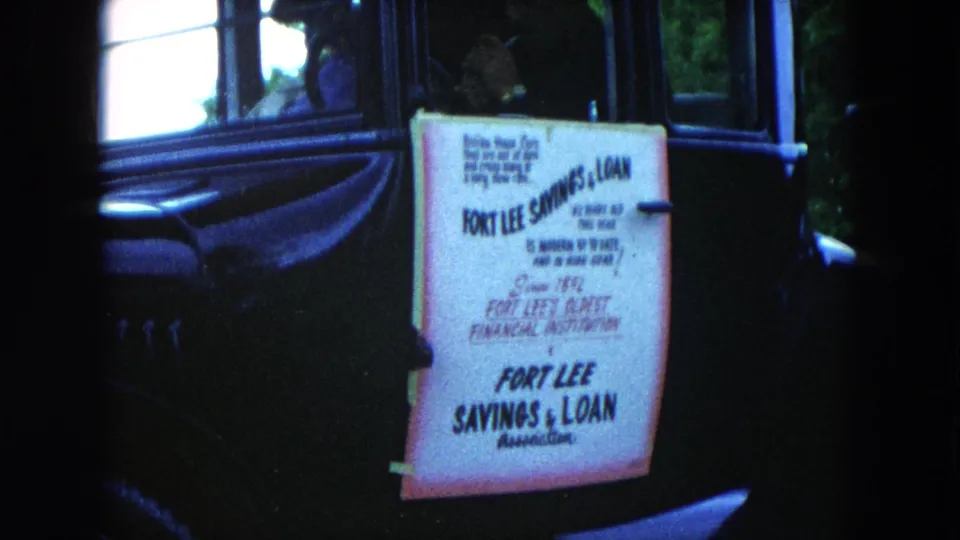 Close 1954 Hicksville frame of a Fort Lee Savings & Loan sign fastened to the side of a black vintage car.