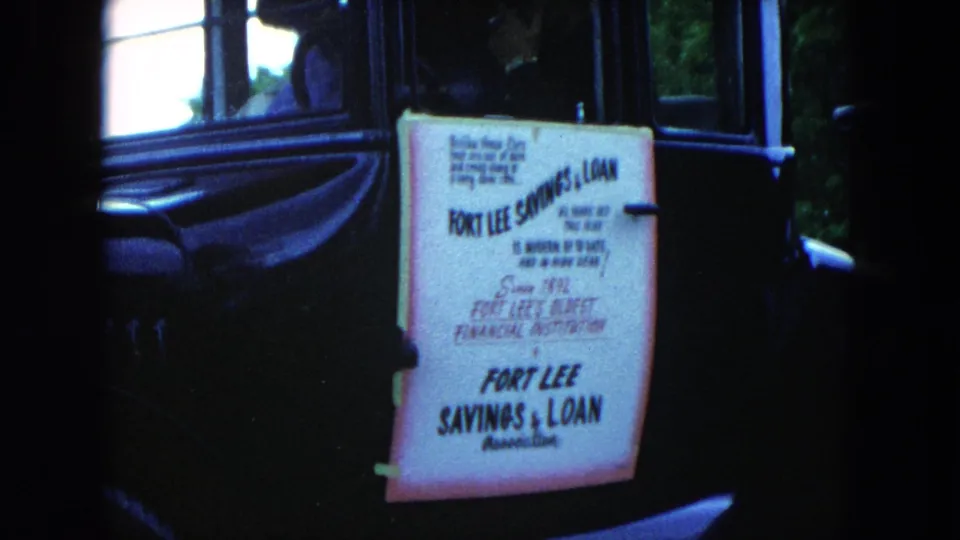 1954 Hicksville frame centered on the Fort Lee Savings & Loan sign on a dark vintage car with people visible inside.