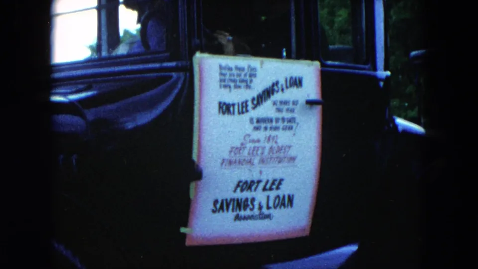 Final 1954 Hicksville frame of the Fort Lee Savings & Loan sign attached to the side of a black vintage car.