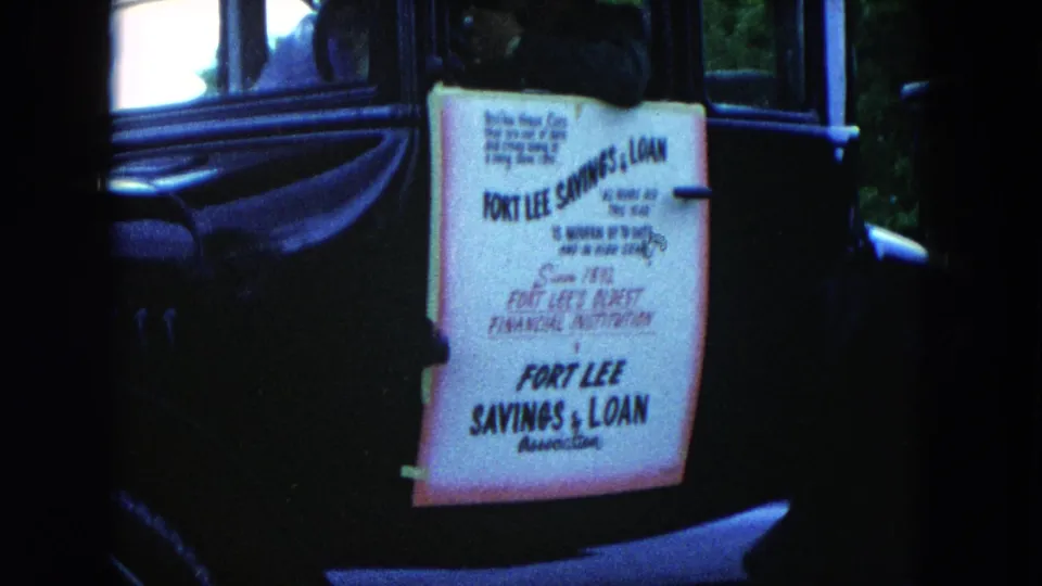 Another close view of the Fort Lee Savings & Loan sign attached to a dark car, with trees visible beyond the vehicle.