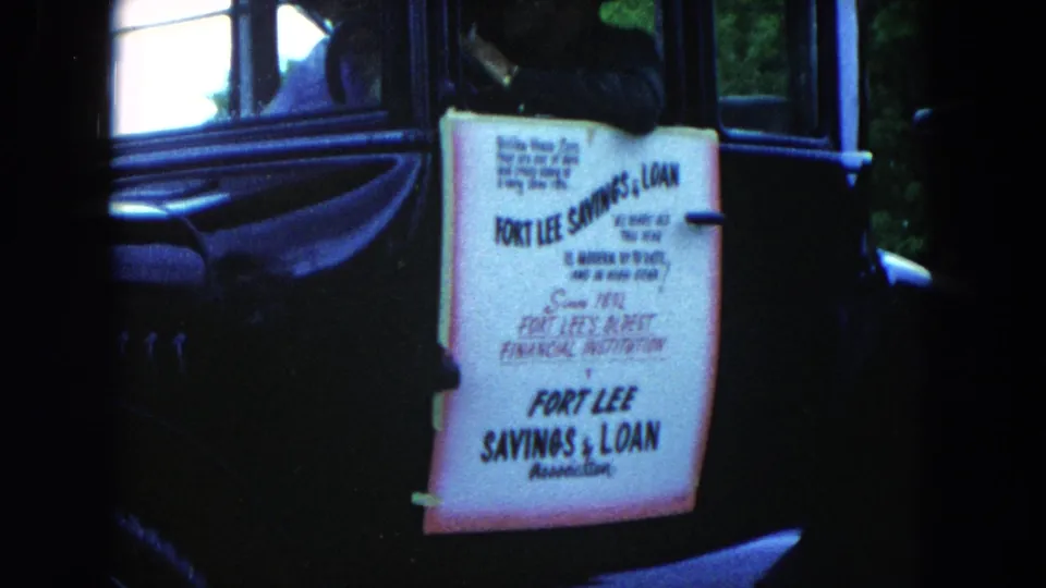 Close-up of the hand-lettered Fort Lee Savings & Loan Association parade sign on a dark car in 1950s Hicksville footage.