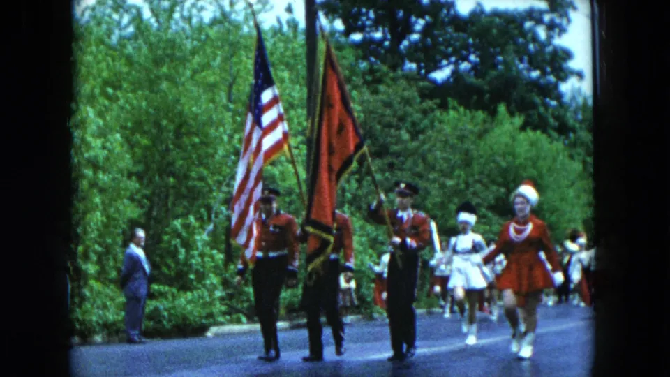 Parade marchers in red and white uniforms carry the American flag and a red banner along a tree-lined road in Hicksville footage.