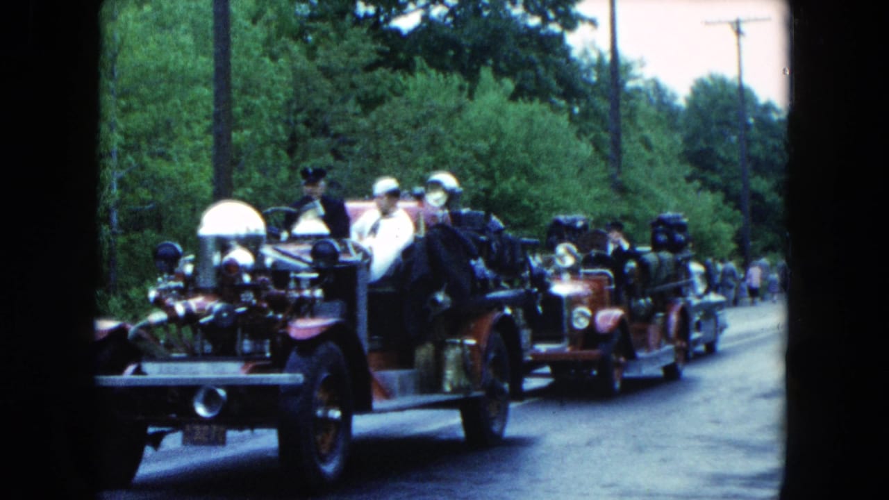 Antique fire engines passing spectators on a tree-lined road.