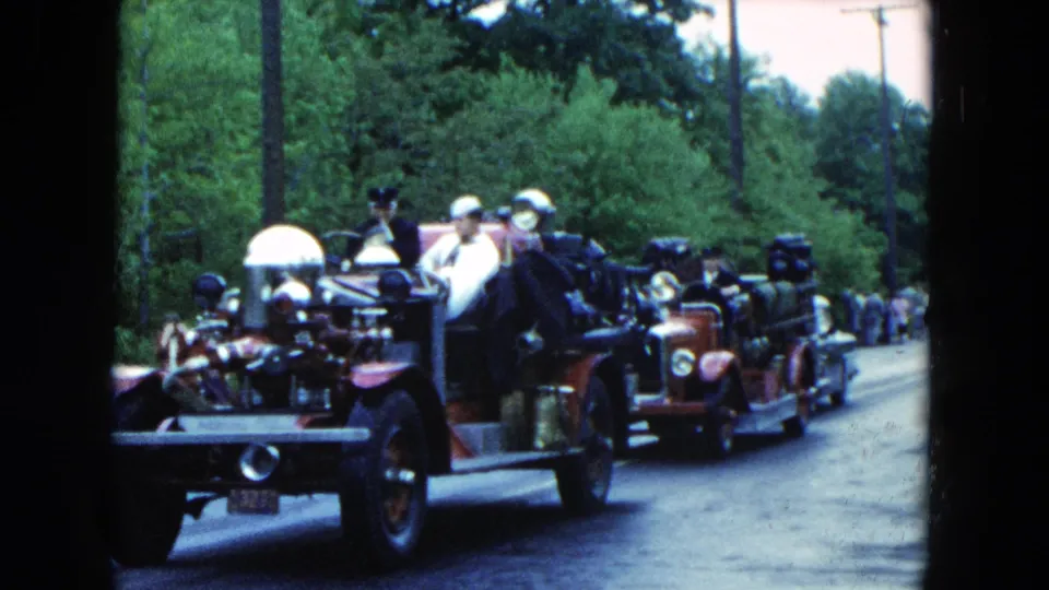 1954 Hicksville parade view with antique fire apparatus, white-helmeted riders, trees, and spectators along the roadway.