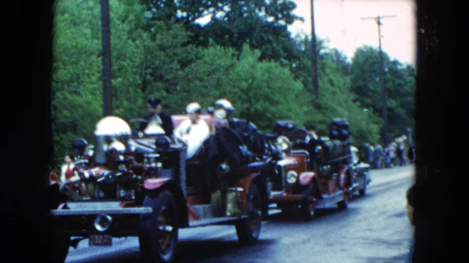 Home-movie frame from Hicksville, New York, showing red fire engines in formation on a tree-lined road in 1954.