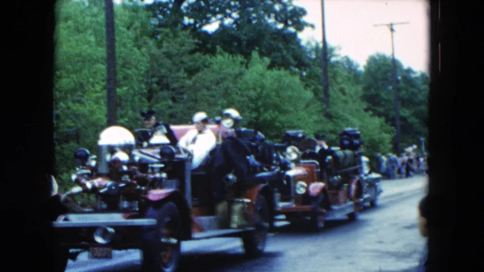 1954 Hicksville parade frame with antique red fire trucks, uniformed riders, spectators, and visible 8mm film borders.