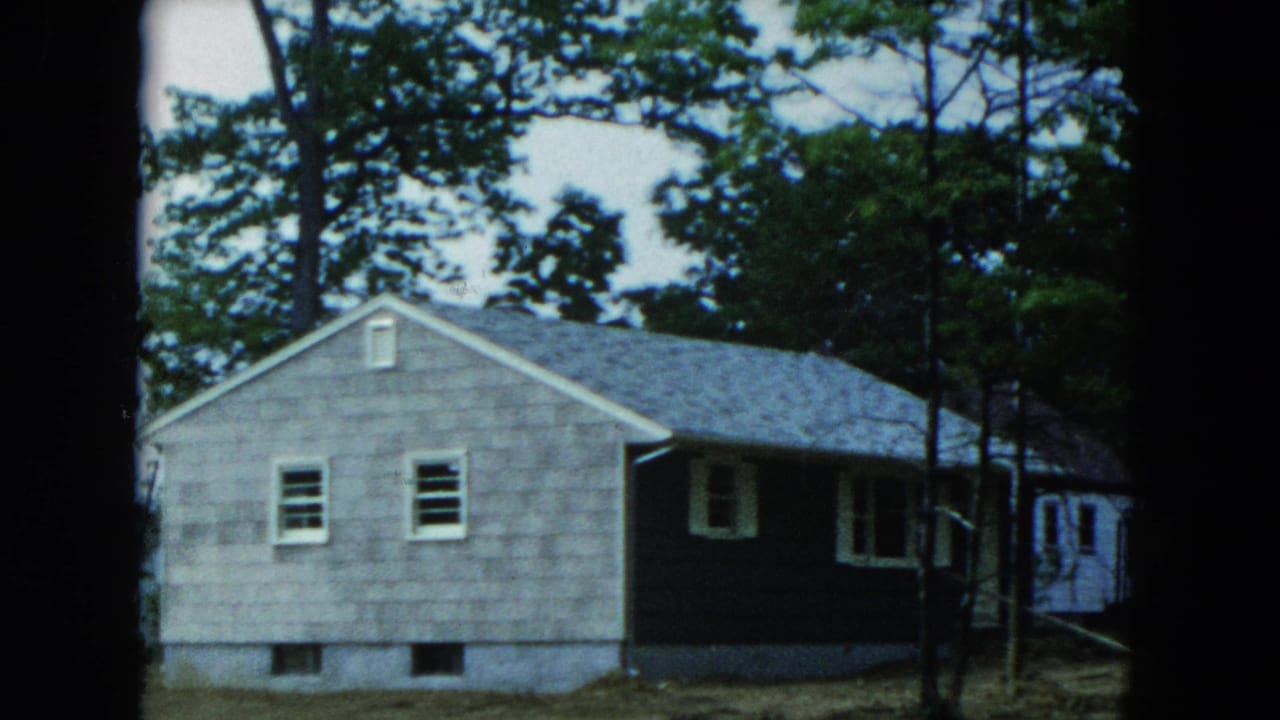 1954 Hicksville home-movie frame showing a grey shingle house with dark trim among tall trees.
