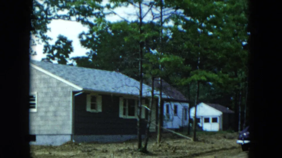 1954 home-movie frame showing the house side, white shed, dirt driveway, and part of a parked car.