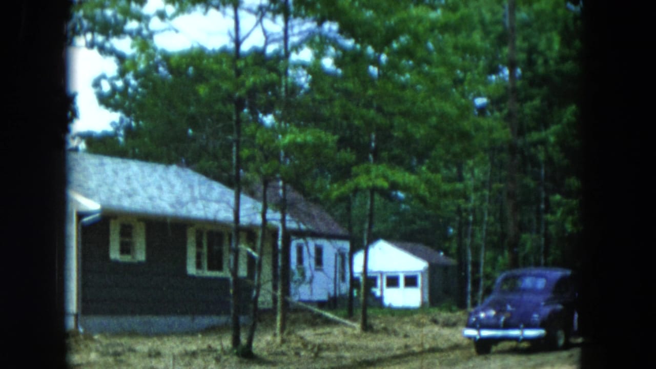 1954 Hicksville, New York home-movie frame showing a dark-sided house, small white shed, tall trees, and a parked mid-century car.