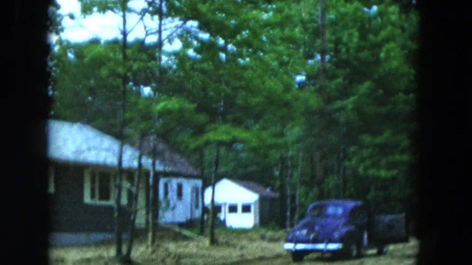 1954 Hicksville home-movie view of a wooded lot with a single-story house, detached white shed, and parked dark automobile.