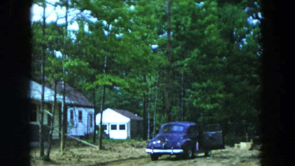 1954 Hicksville frame showing a dark parked automobile with an open door near wooded houses and a small white shed.