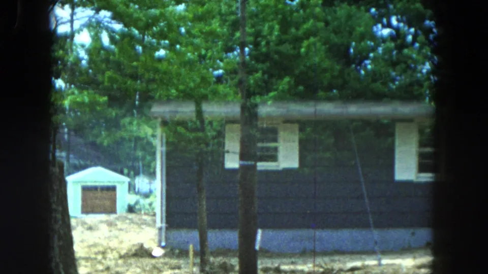 1954 Hicksville home-movie frame with a closer view of a dark-sided house, white trim, trees, and a small outbuilding behind it.