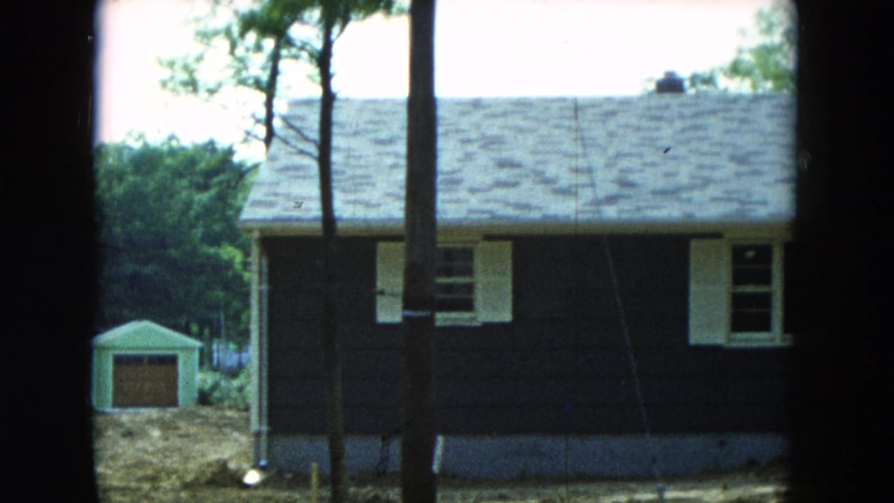 1954 Hicksville, New York home-movie frame showing a dark-sided house with white trim, trees in front, and a small yard shed.