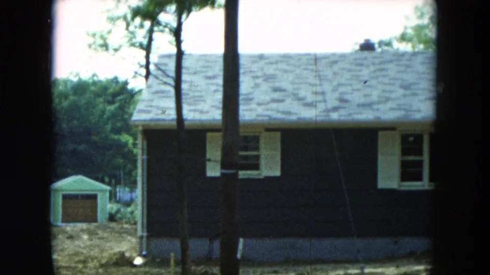 1954 Hicksville, New York home-movie frame showing a dark-sided house with white trim, trees in front, and a small yard shed.