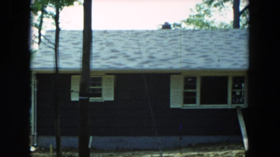 1954 Hicksville, New York frame of a modest dark-sided house with white shutters, shingled roof, and tall trees crossing the view.