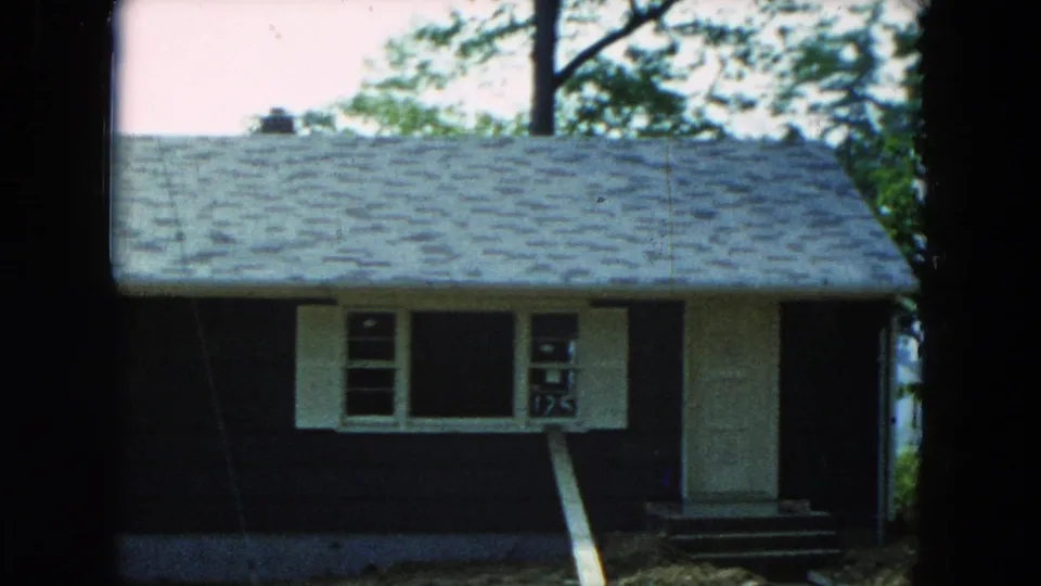 1954 Hicksville, New York frame showing the house entrance area, shuttered windows, dark siding, and trees around the lot.