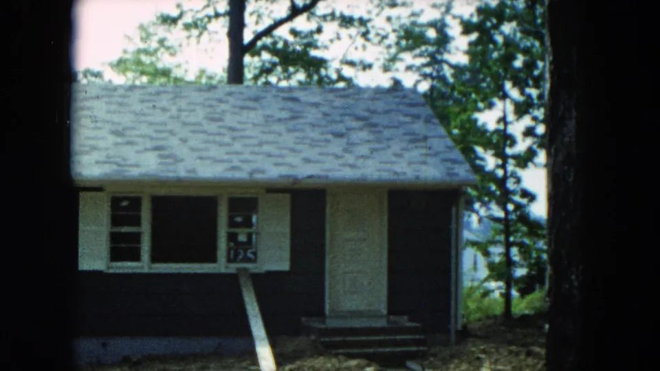 1954 Hicksville exterior view of a small residential house with white trim, porch steps, surrounding trees, and a board leaning near the window.