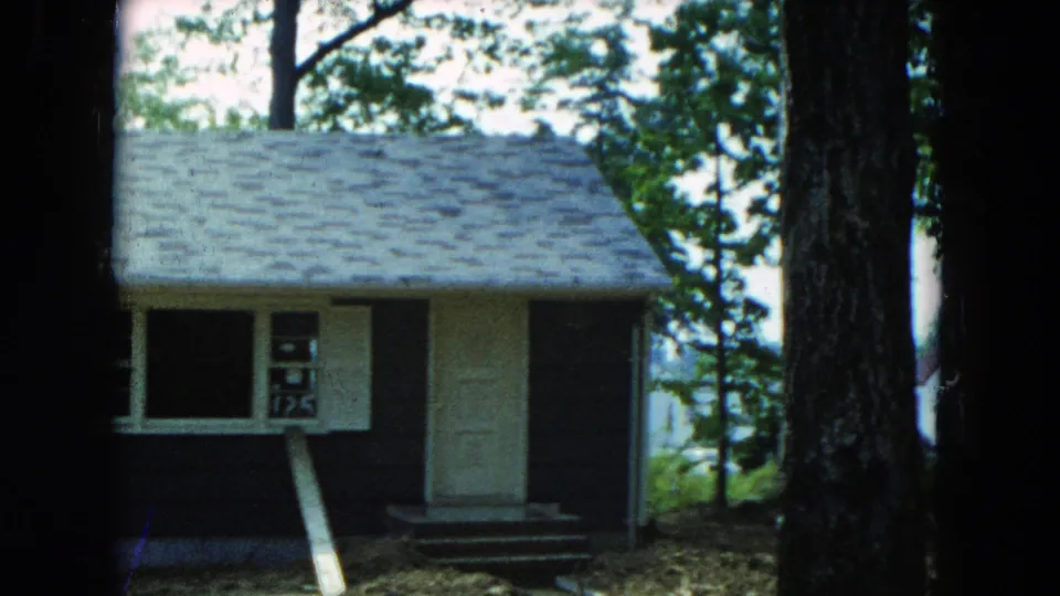 1954 Hicksville home-movie frame with a closer view of the house facade, roofline, white door, dark siding, and a large tree trunk nearby.