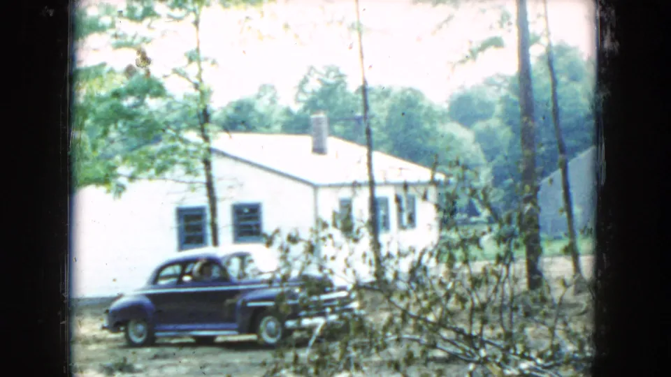 1954 Hicksville frame showing a dark mid-century sedan parked near a white residential house, seen through foreground foliage.