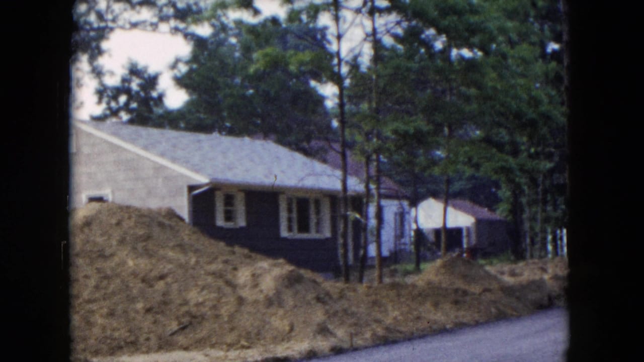 1954 Hicksville, New York home-movie frame showing a brown house, white trim, pine trees, earth piles, and a paved road edge.