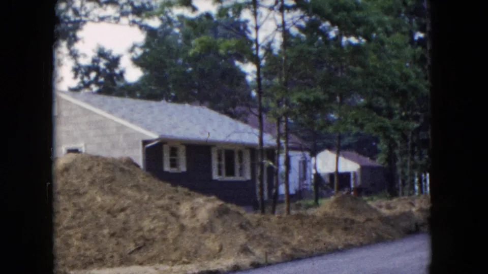 1954 Hicksville, New York home-movie frame showing a brown house, white trim, pine trees, earth piles, and a paved road edge.