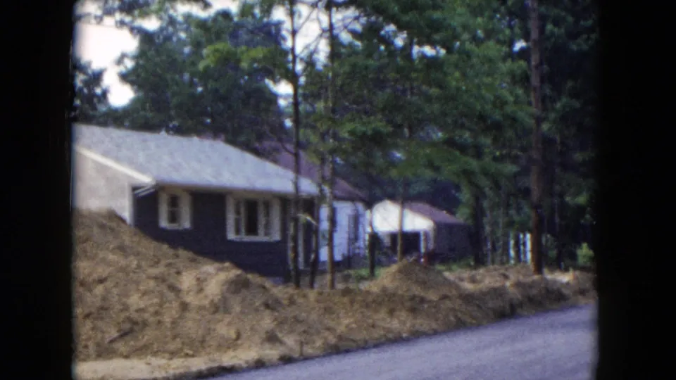 1954 Hicksville residential exterior with a dark house, white trim, pine trees, earth piles, and a quiet paved street.