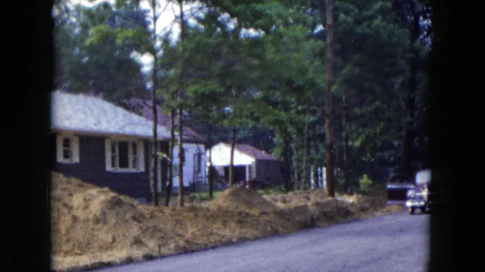 Hicksville, New York 1954 home-movie still showing a house, roadside earth piles, trees, and a mid-century car at the road edge.