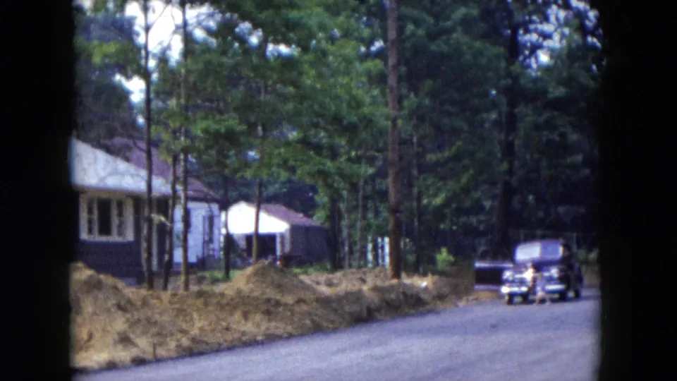 1954 Hicksville home-movie frame with a car on a paved residential road, a brown house, white trim, trees, and construction dirt piles.
