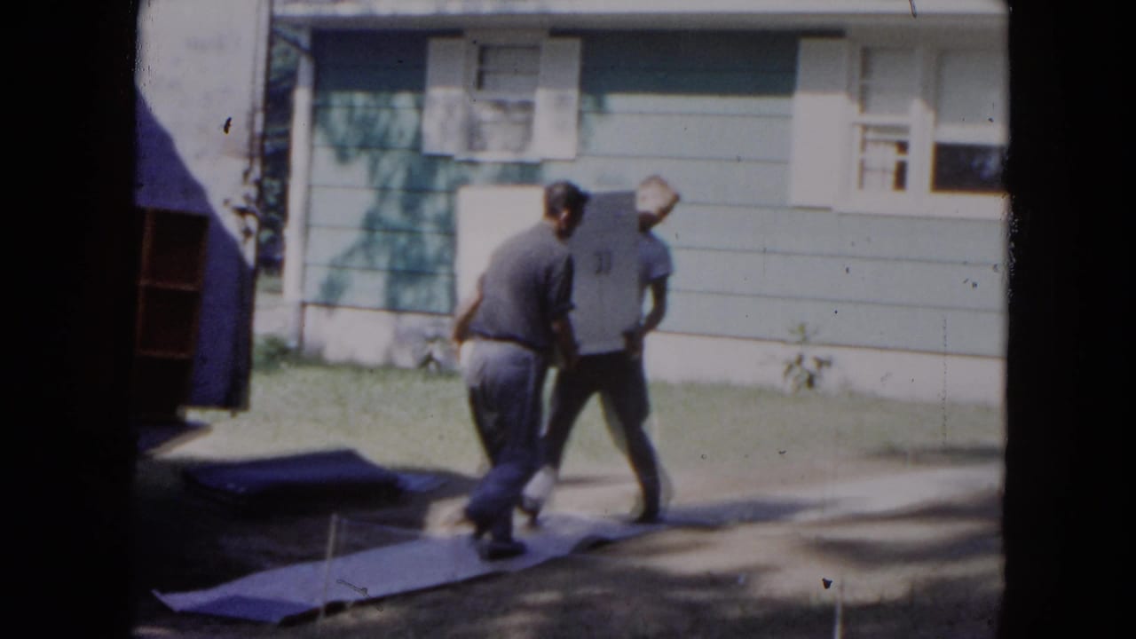 Two men carry a large white appliance across a walkway beside a blue house.