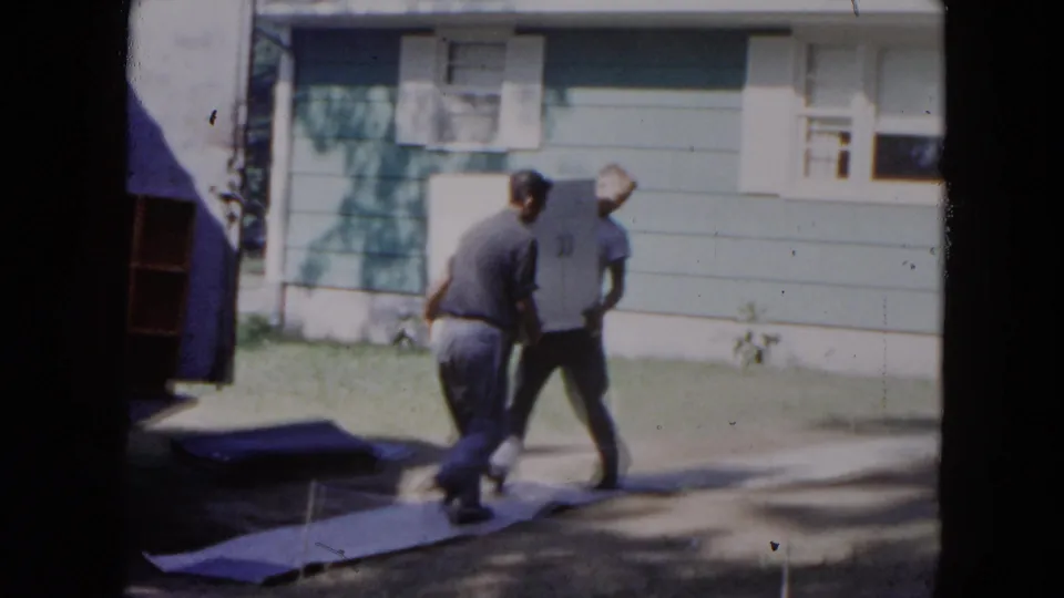 In Hicksville, New York, 1954, two adult men carry a large white appliance past a blue-sided suburban house.