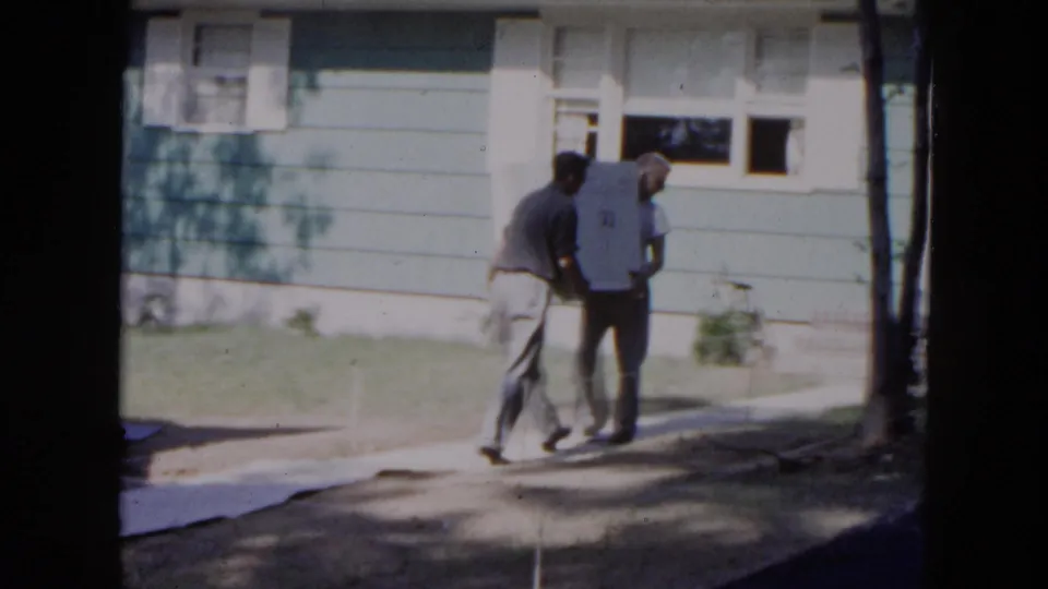 The men carry the white appliance closer to the entrance of a Hicksville suburban house in 1954 home-movie footage.