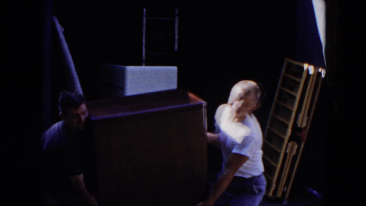In 1954 Hicksville home-movie footage, adult men maneuver a large furniture piece through a shadowed doorway area.
