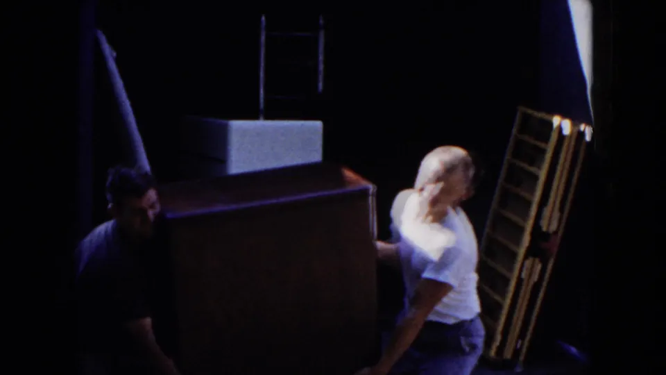 In 1954 Hicksville home-movie footage, adult men maneuver a large furniture piece through a shadowed doorway area.